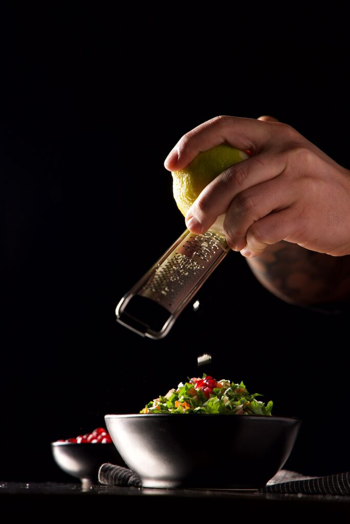 A chef grating fresh lemon zest over a bowl of tabouleh, garnished with pomegranate seeds.