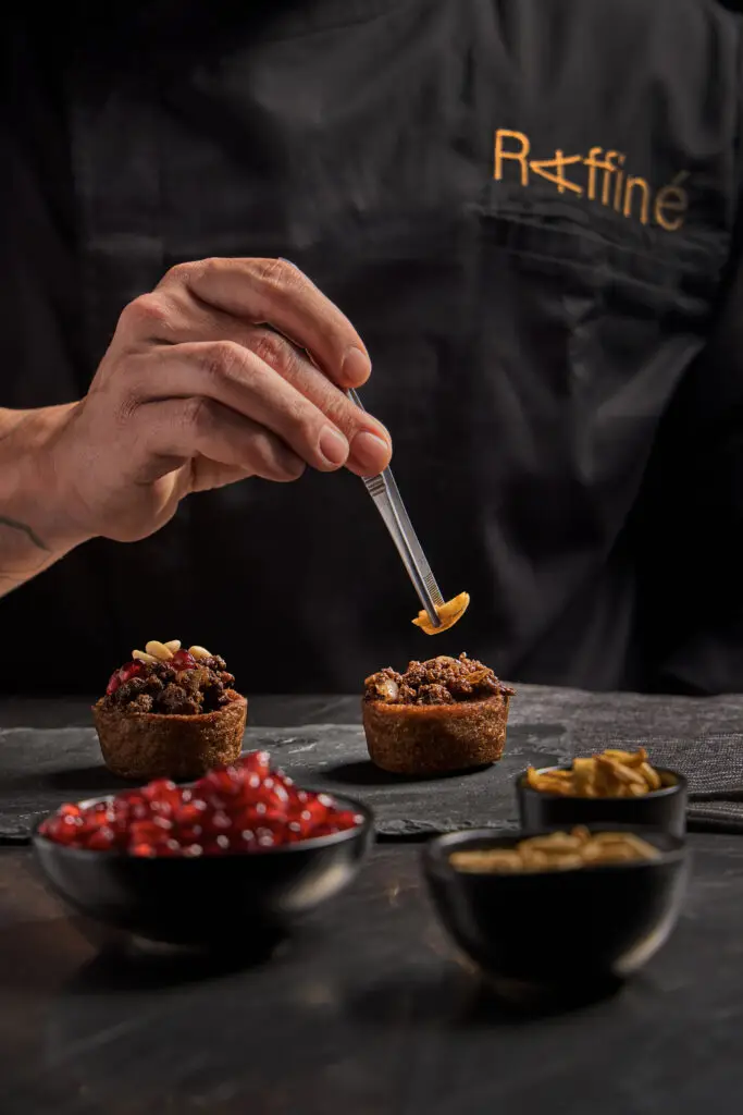 Chef placing almond on open-faced kibbeh cupcake with tweezers at Raffiné Lebanese Restaurant & Wine Bar, surrounded by pomegranate seeds and nuts.